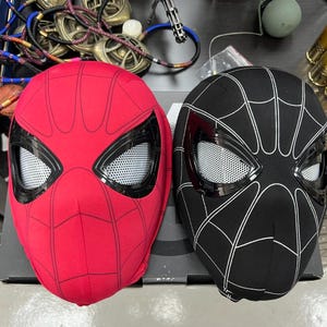 Op de afbeelding: Twee Spider-Man maskers, een rode en een zwarte, met witte spinnenwebben en gaasoogbedekkingen.