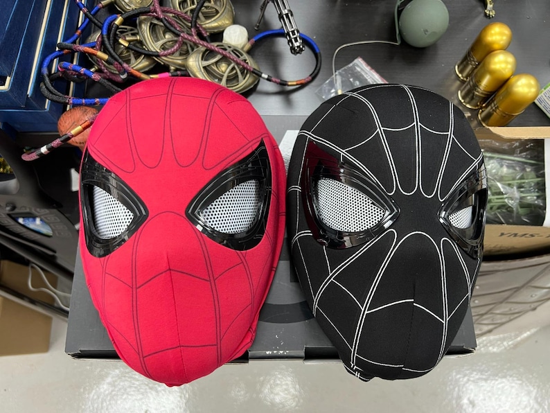 Op de afbeelding: Twee Spider-Man maskers, een rode en een zwarte, met witte spinnenwebben en gaasoogbedekkingen.