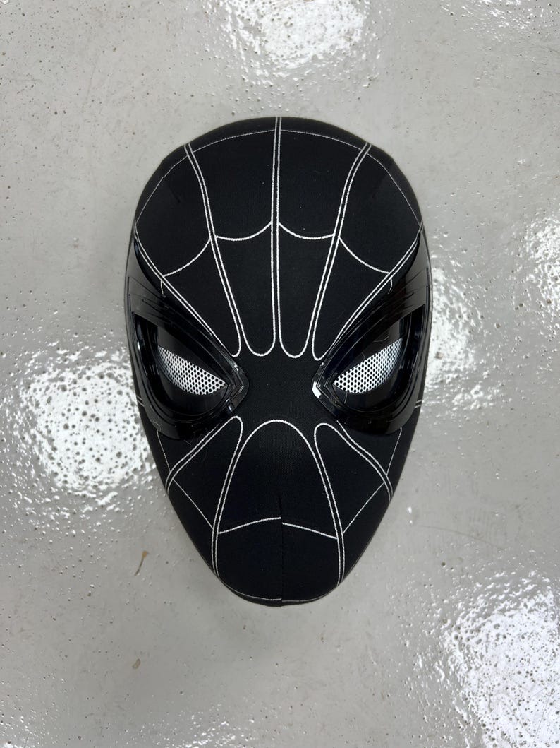 Op de afbeelding: Zwart-witte Spider-Man-masker met een zwart gaas dat de ogen bedekt. Het masker is gemaakt van stof en heeft een 3D-ontwerp.