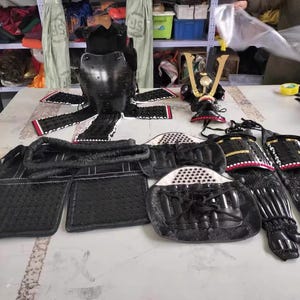 May include: A set of black samurai armor, including a helmet, chest plate, and leg guards. The armor is made of leather and metal and has intricate details. The armor is displayed on a table with a white background.