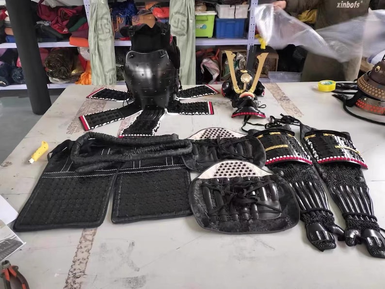 May include: A set of black samurai armor, including a helmet, chest plate, and leg guards. The armor is made of leather and metal and has intricate details. The armor is displayed on a table with a white background.