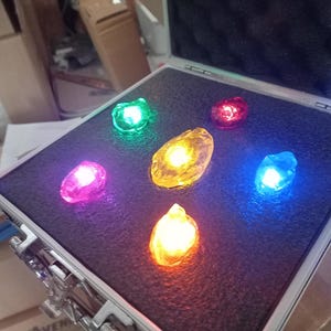 Puede incluir: Una caja negra con seis gemas de colores en su interior. Las gemas están dispuestas en círculo y se iluminan desde el interior. Las gemas son roja, amarilla, verde, azul, morada y naranja.