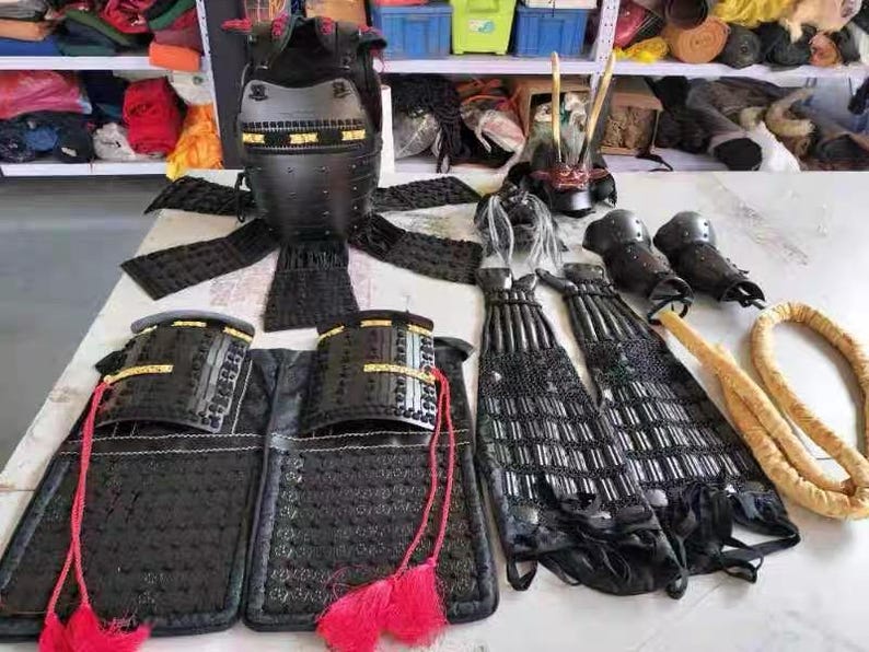 May include: A black samurai armor set with a helmet, chest plate, arm guards, and leg guards. The armor is made of metal and has a traditional Japanese design.