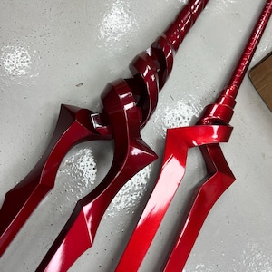 May include: Two red metal swords with intricate designs. The swords have a shiny, polished finish and are displayed on a white surface.