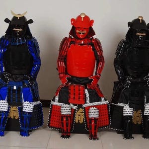 May include: Three samurai armor figures, one in red, one in blue, and one in black. Each figure is wearing a helmet and armor with intricate details. The figures are standing on a white surface.