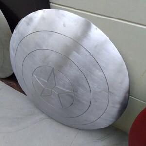May include: A silver-colored Captain America shield with a five-pointed star in the center. The shield has concentric circles around the star.