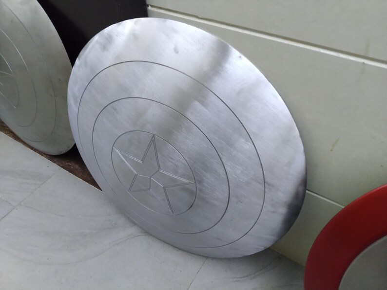 May include: A silver-colored Captain America shield with a five-pointed star in the center. The shield has concentric circles around the star.