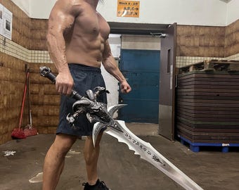 May include: A large, ornate fantasy sword with a silver blade and a detailed black handle. The sword features intricate carvings and a stylized demon head at the hilt. The blade has etched inscriptions. The person is wearing black shorts and black Puma sneakers.