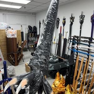 Könnte beinhalten: Ein großes, schwarz-graues Fantasy-Schwert mit strukturierter Klinge und klauenartigem Griff. Das Schwert hat eine scharfe, spitze Spitze und wird vor einer Ausstellung anderer Schwerter und Waffen gehalten.