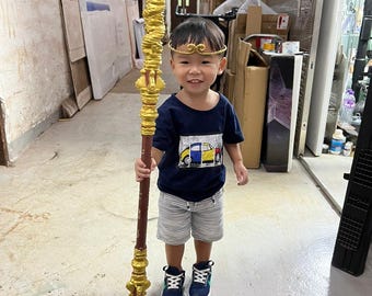 May include: A young child wearing a blue shirt and grey shorts holds a long, gold-painted staff with a brown wooden base. The staff has intricate details and is decorated with gold accents. The child is smiling and looking at the camera.