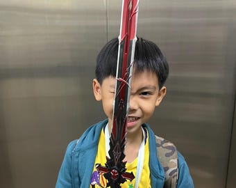 May include: A young person holds a red and black sword with a cross-shaped handle. The sword is held in front of the person's face, partially obscuring their features. The person is wearing a blue jacket over a yellow shirt with a cartoon design.