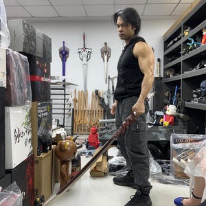 Könnte beinhalten: Ein Mann in einem schwarzen ärmellosen Hemd und schwarzen Hosen hält ein langes, rotgriffiges Schwert mit einer silbernen Klinge. Er steht in einem Raum mit Regalen voller Schwerter und anderer Waffen.