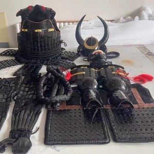 May include: A black samurai armor set with a horned helmet, chest plate, and arm guards. The armor is made of metal and has a textured finish. The set also includes a pair of black leggings and a black fabric sash.