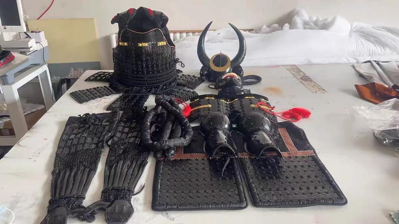 May include: A black samurai armor set with a horned helmet, chest plate, and arm guards. The armor is made of metal and has a textured finish. The set also includes a pair of black leggings and a black fabric sash.