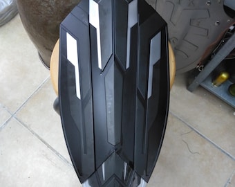 May include: A black and silver shield with a geometric design. The shield is made of a hard plastic material and has a raised edge around the perimeter.