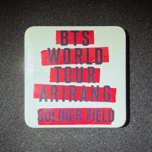 May include: A square, off-white sticker with the text "BTS WORLD TOUR ARIRANG SOLDIER FIELD" in red and black lettering. The text is arranged in horizontal lines, with the words "BTS", "WORLD TOUR", "ARIRANG", and "SOLDIER FIELD" prominently displayed.