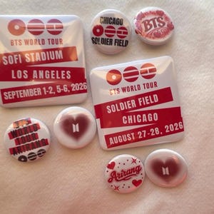 May include: A collection of white and red BTS World Tour buttons and pins. The pins feature text such as "SOFI STADIUM LOS ANGELES SEPTEMBER 1-2, 5-6, 2026" and "SOLDIER FIELD CHICAGO AUGUST 27-28, 2026". Some pins have heart designs.