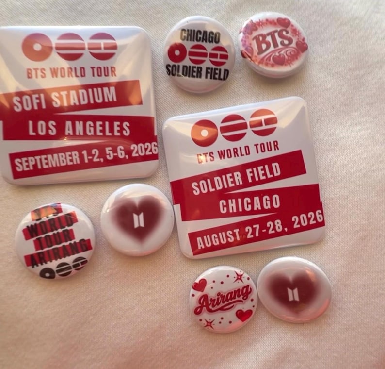 May include: A collection of white and red BTS World Tour buttons and pins. The pins feature text such as "SOFI STADIUM LOS ANGELES SEPTEMBER 1-2, 5-6, 2026" and "SOLDIER FIELD CHICAGO AUGUST 27-28, 2026". Some pins have heart designs.