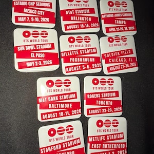 May include: A collection of white and red BTS World Tour souvenir pins. Each pin features the tour logo and the name of a stadium, city, and dates for the 2026 tour, including locations like Mexico City and Arlington.