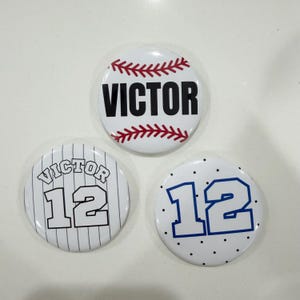 May include: Three white baseball-themed buttons. One button has the name "VICTOR" in black with red stitching. Another has "VICTOR" and the number "12" in a baseball jersey style. The third button has the number "12" in blue.