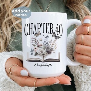 May include: White ceramic mug with a black and white design of a book with flowers and butterflies. The text "Chapter 40" is printed on the mug. The mug is being held by a person.