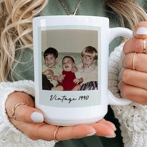 Puede incluir: Taza de cerámica blanca con una fotografía vintage de tres niños. La fotografía es de estilo Polaroid con el texto "Vintage 1990" impreso debajo. La taza la sostiene una persona que lleva una camisa verde y un suéter blanco.