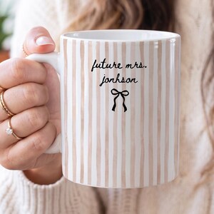 Puede incluir: Una taza de cerámica blanca con rayas verticales de color marrón claro estilo acuarela. El frente de la taza muestra el texto negro 'future mrs. jonhson' en letra cursiva, colocado sobre un pequeño icono de lazo dibujado en negro.