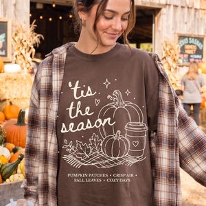 Puede incluir: Una camiseta marrón oscuro con un gráfico de líneas blancas de calabazas, una taza de café y hojas con el texto 'tis the season' y 'PUMPKIN PATCHES • CRISP AIR • FALL LEAVES • COZY DAYS' usada debajo de una camisa de cuadros marrón y blanca.