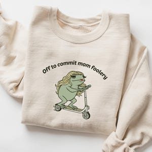 Puede incluir: Sudadera de color arena con el texto "Off to commit mom foolery" sobre una rana de dibujos animados montada en un patinete. La rana tiene pelo largo rubio y pintalabios rojo. La sudadera tiene mangas largas.