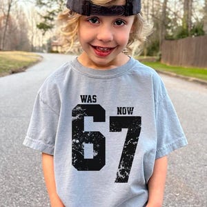 Peut inclure: Un t-shirt gris clair &agrave; manches courtes avec une impression graphique noire vieillie. Le motif comprend le mot WAS au-dessus du nombre 67 et le mot NOW &agrave; droite du nombre. Le t-shirt est port&eacute; avec une casquette de baseball gris fonc&eacute;.
