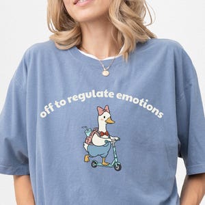 Puede incluir: Camiseta azul vaquero con el texto "off to regulate emotions". La camiseta presenta un dibujo de un ganso de dibujos animados montado en un patinete. El ganso lleva un lazo y una mochila. La camiseta es de manga corta y cuello redondo.