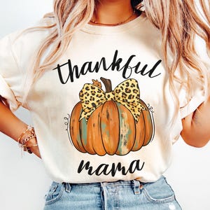 Puede incluir: Camiseta color marfil con las palabras "thankful mama" en escritura negra. Una gran calabaza naranja con un lazo con estampado de leopardo está centrada en la camiseta. La camiseta es de un material suave.