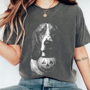 Puede incluir: Una camiseta gris carbón de manga corta con un estampado gráfico en blanco y negro de un perro sabueso de orejas largas. El perro está situado sobre un cubo de calabaza jack-o'-lantern tallado con una cara sonriente.
