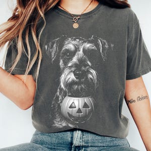 May include: A charcoal gray short-sleeve t-shirt featuring a black and white graphic print of a Schnauzer dog holding a carved pumpkin jack-o'-lantern in its mouth. The shirt has a crew neckline and a relaxed fit, showcasing a detailed illustration.