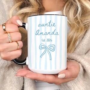 Puede incluir: Taza de cerámica blanca con asa negra, con rayas verticales azul claro y un diseño de lazo. El texto "auntie Amanda Est. 2026" está impreso en una fuente azul claro. La taza se sostiene frente a un suéter de tono neutro.