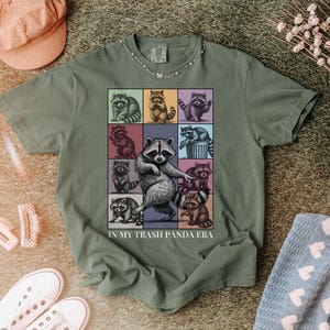 May include: Moss green t-shirt featuring a colorful graphic of raccoons in various poses. The phrase "IN MY TRASH PANDA ERA" is printed below the graphic. The shirt is styled with a pink cap, white sneakers, and a blue patterned towel.