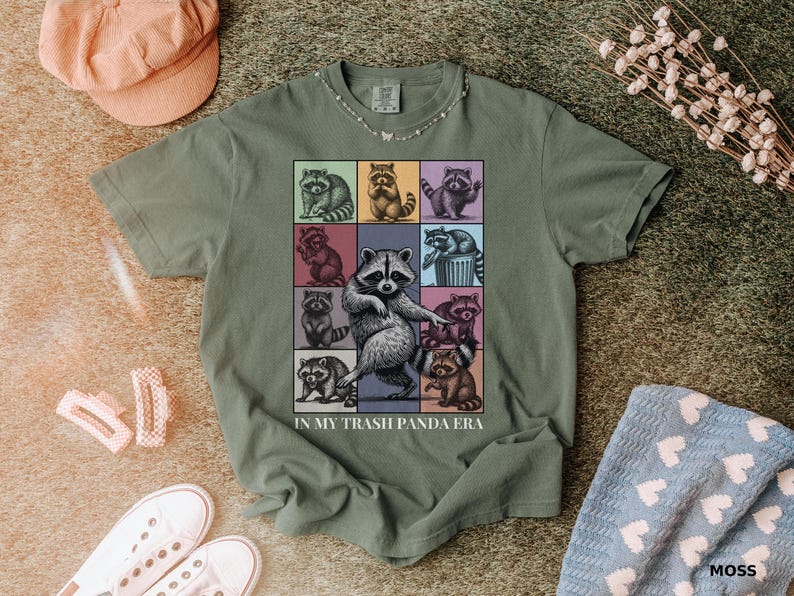 May include: Moss green t-shirt featuring a colorful graphic of raccoons in various poses. The phrase "IN MY TRASH PANDA ERA" is printed below the graphic. The shirt is styled with a pink cap, white sneakers, and a blue patterned towel.