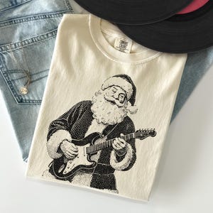 Puede incluir: Camiseta color crema con una ilustración en blanco y negro de Papá Noel tocando la guitarra eléctrica. La camiseta está doblada sobre unos vaqueros azul claro y dos discos de vinilo.