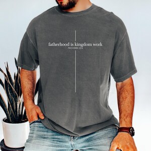 May include: A dark gray t-shirt with the text "fatherhood is kingdom work" and the Bible verse Proverbs 22:6 printed in white. A vertical white line forms a cross. The shirt is worn with blue jeans.