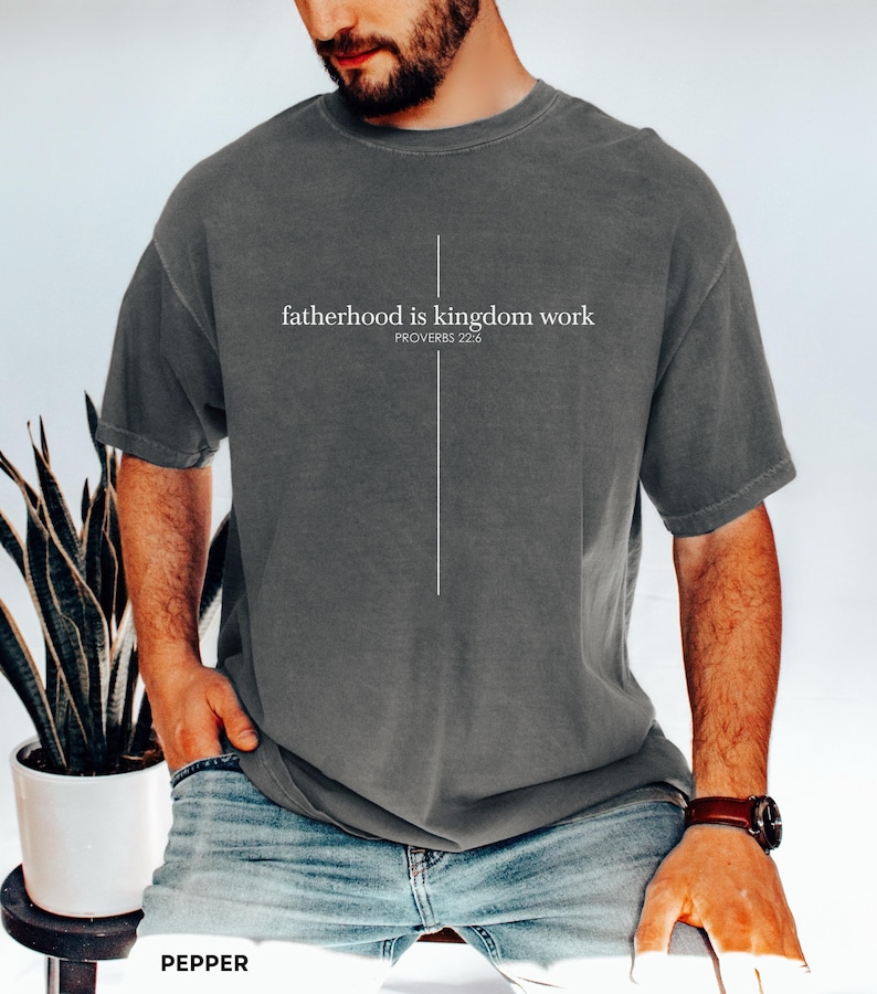May include: A dark gray t-shirt with the text "fatherhood is kingdom work" and the Bible verse Proverbs 22:6 printed in white. A vertical white line forms a cross. The shirt is worn with blue jeans.