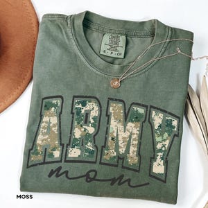 Puede incluir: Camiseta verde musgo con las palabras "ARMY MOM" en un diseño de camuflaje. La camiseta tiene cuello redondo y mangas cortas. Un collar dorado con un colgante está drapeado sobre la camiseta. La camiseta está doblada sobre una superficie blanca.