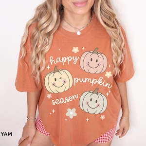 Puede incluir: Una camiseta de manga corta color naranja quemado con tres calabazas de colores claros con caras sonrientes y el texto "happy pumpkin season" en cursiva blanca, acentuado con pequeños iconos de flores y estrellas blancas al frente.