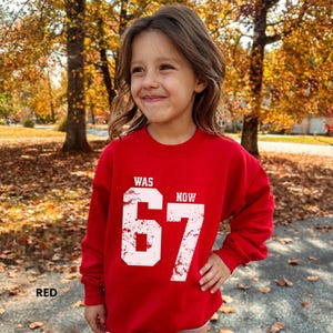 Peut inclure: Un sweat-shirt rouge &agrave; manches longues avec un texte blanc au style vieilli indiquant WAS NOW 67. Le motif est centr&eacute; sur la poitrine avec le nombre 67 dans une grande police us&eacute;e, tandis que les mots WAS et NOW sont plus petits.