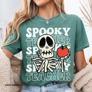 Puede incluir: Camiseta verde claro con el texto blanco "SPOOKY TEACHER", apilado verticalmente. Un gráfico de esqueleto sostiene una manzana roja. La camiseta es de cuello redondo y tiene mangas cortas. Las palabras "LIGHT GREEN" están abajo a la izquierda.