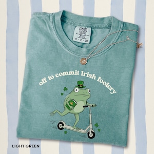 Puede incluir: Camiseta verde claro con el texto "off to commit Irish foolery" y una rana de dibujos animados con un sombrero de duende montada en un patinete. Un collar de oro con un colgante está sobre la camiseta.