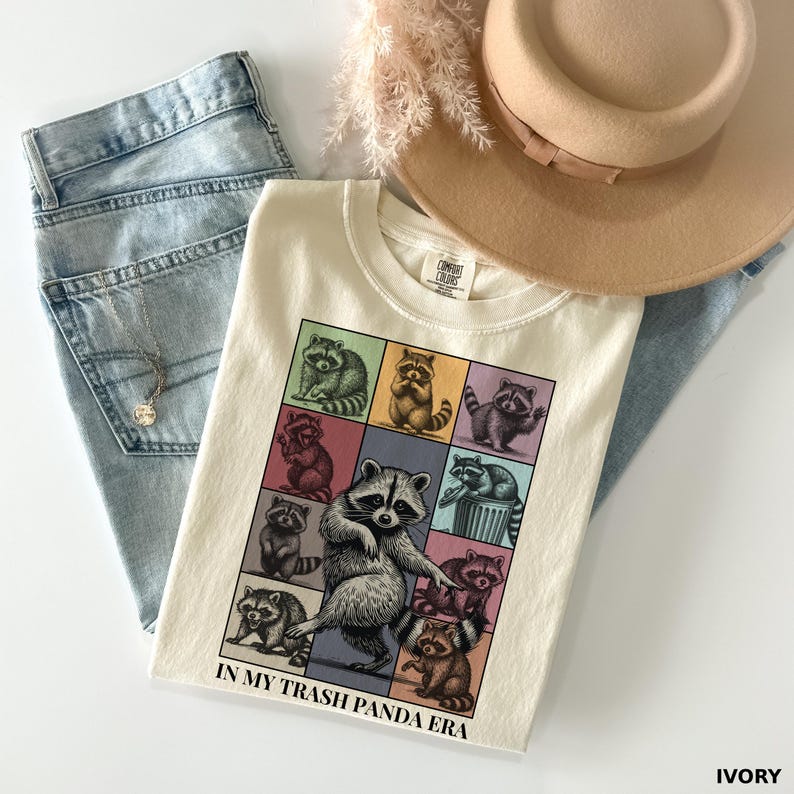 May include: Ivory-colored t-shirt with a colorful raccoon graphic and the text "IN MY TRASH PANDA ERA." The shirt is paired with light blue jeans, a gold necklace, and a tan hat. The shirt is made of a soft, comfortable material.