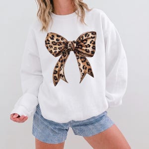 Puede incluir: Sudadera blanca con un gran diseño de lazo con estampado de leopardo. El lazo tiene manchas marrones y negras. Se combina con pantalones cortos a rayas azules y blancas. El estilo es casual y femenino.