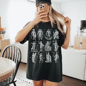 May include: Black t-shirt featuring a white illustration of various mythological figures. The shirt is worn with a black baseball cap that reads "COWBOY HAT". The person is holding a phone in front of their face.