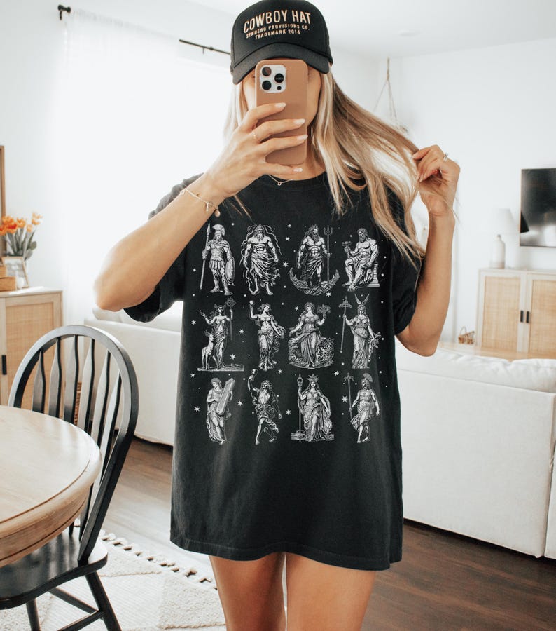 May include: Black t-shirt featuring a white illustration of various mythological figures. The shirt is worn with a black baseball cap that reads "COWBOY HAT". The person is holding a phone in front of their face.