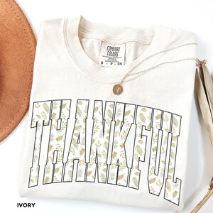 May include: Ivory-colored t-shirt with the word "THANKFUL" in large, block letters filled with a floral pattern of green and gold leaves. A gold necklace with a pendant rests on the shirt. The shirt is folded.